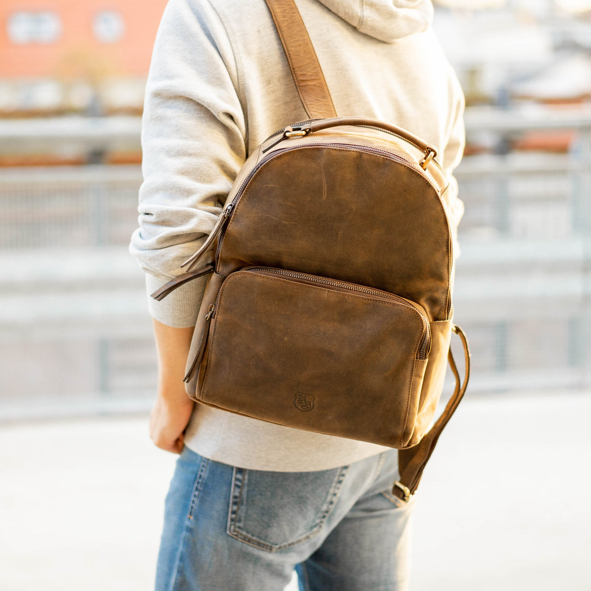 Person trägt einen braunen Lederrucksack mit Reißverschluss-Hauptfach und Vortasche über der Schulter, Büffelleder mit Wappenprägung – Lederrucksack kaufen